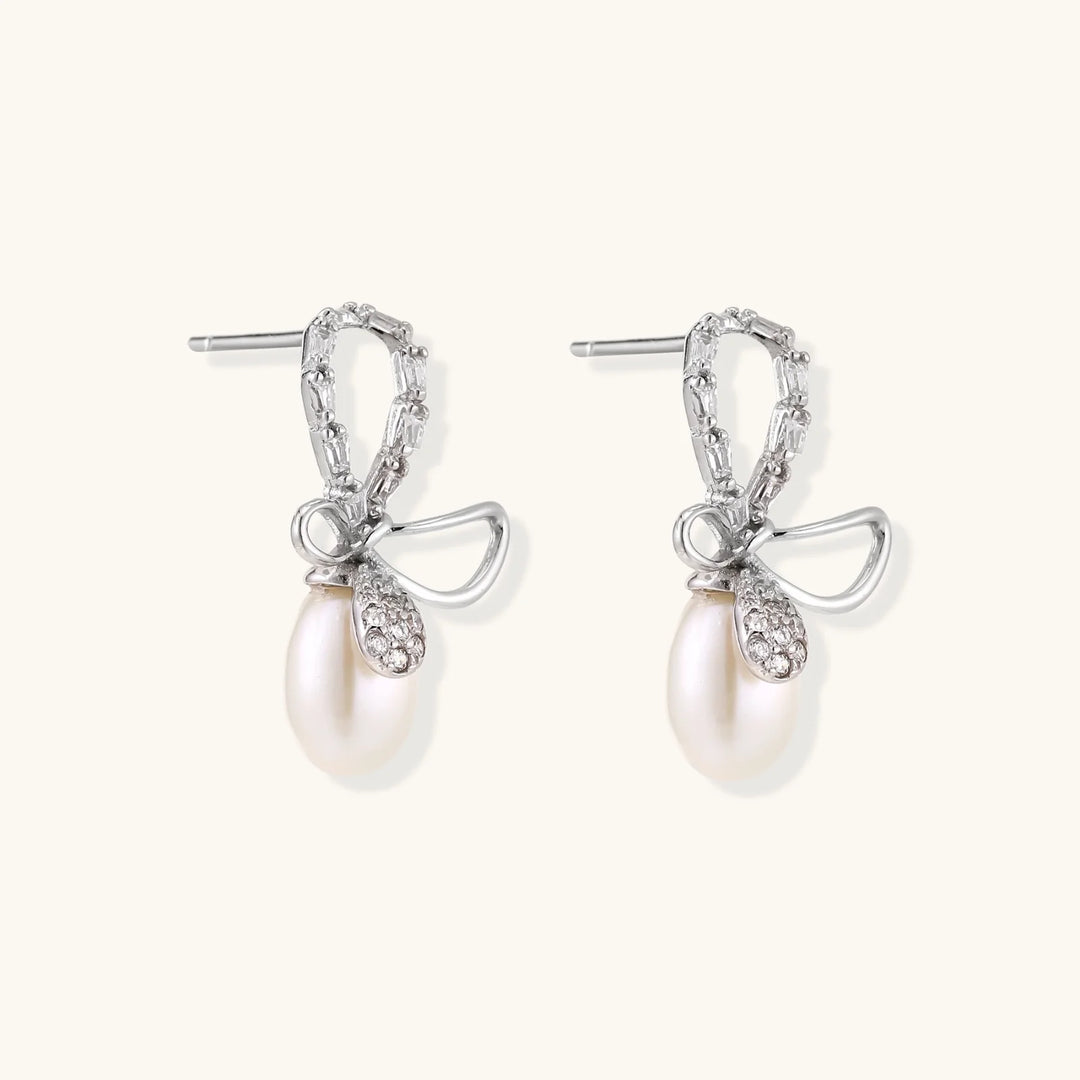 Silver bow-shaped stud earrings with baguette crystals and dangling white freshwater pearls on cream background