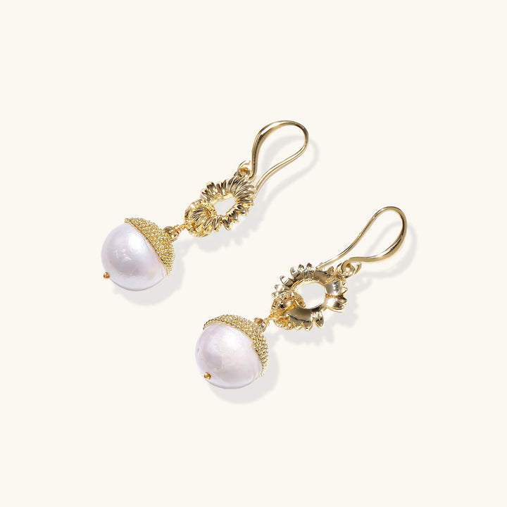 Gold sunburst charm earrings with baroque pearl drops on French hooks against cream background