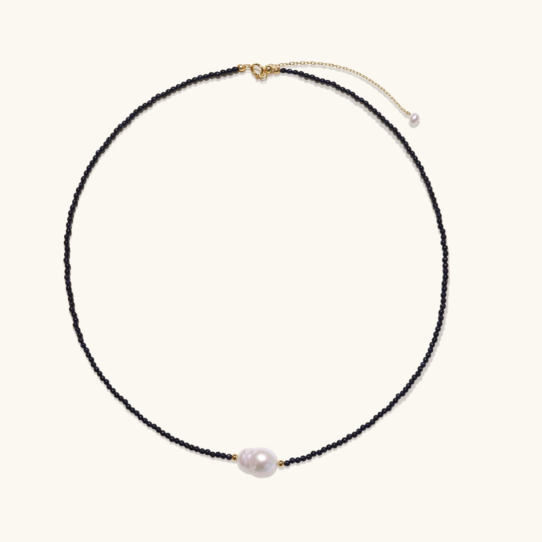 Delicate necklace with black blue sandstone beads and baroque pearl center on white background with gold accents.