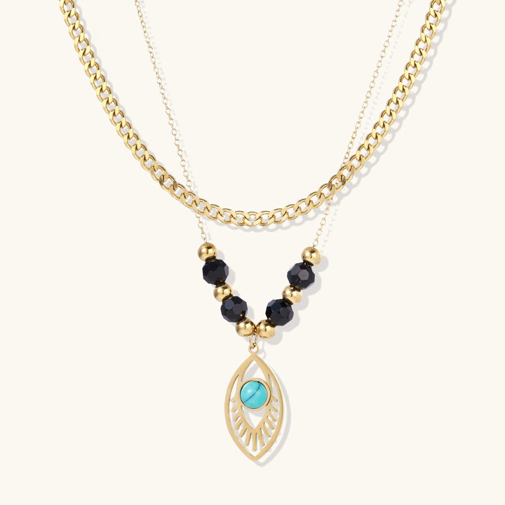 Layered gold necklace with turquoise evil eye pendant and black quartz beads on cream background